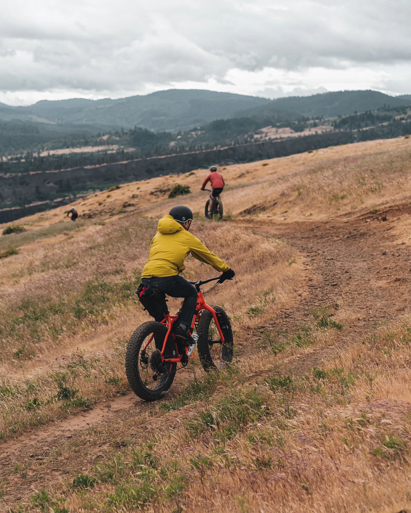 A rider in a yellow jacket on a fat bike descending a grassy hillside trail