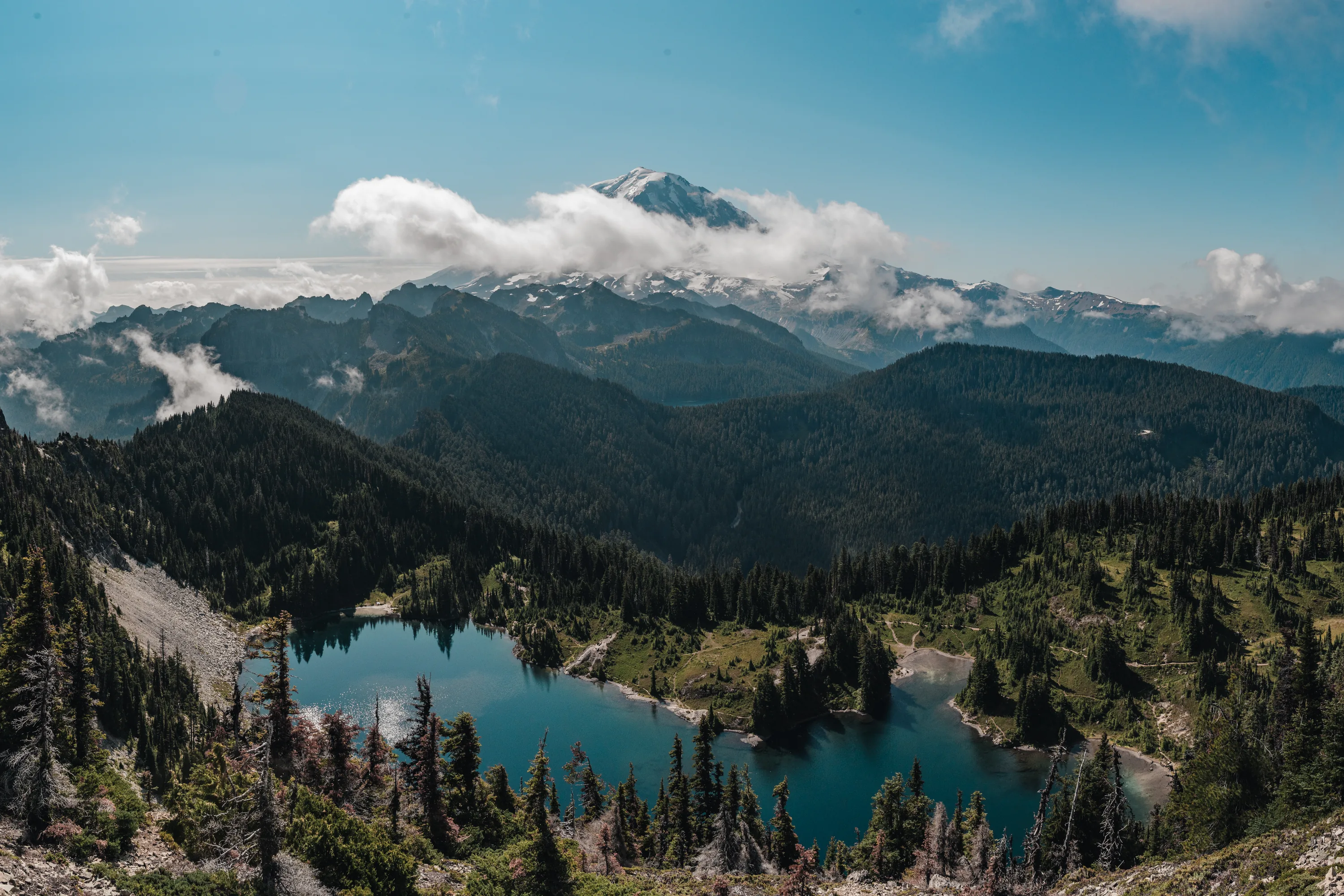 A snow-capped peak above clouds, with a blue alpine lake in a forested valley below